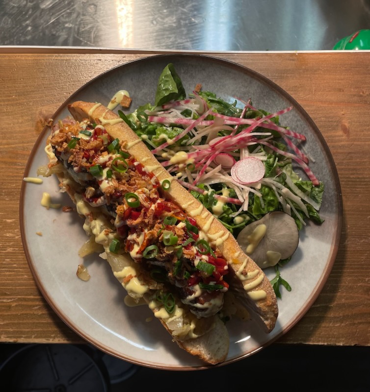 Hot‑dog maison : compotée de fenouil, poivrons grillés, pain croustillant, cébette & oignons frits, saucisse artisanale grillée